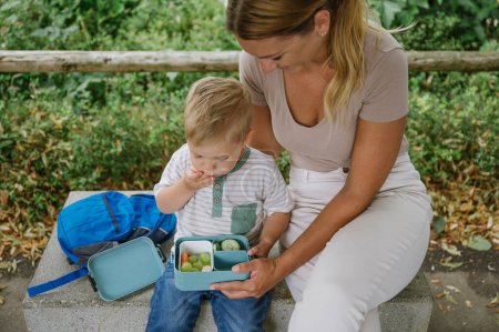 Téléchargez les photos : Culture mère en vêtements décontractés assis sur le banc à côté du petit garçon de la maternelle et manger des légumes frais pour le déjeuner tout en passant du temps ensemble après l'école le jour ensoleillé - en image libre de droit