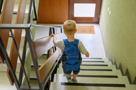 Téléchargez les photos : Vue arrière du petit garçon aux cheveux blonds en vêtements décontractés avec sac à dos debout sur les escaliers avant d'aller à la maternelle le matin. concept de famille - en image libre de droit