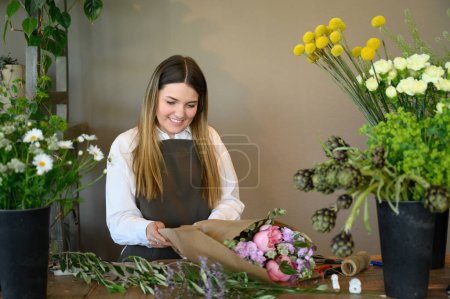 Photo for Young cheerful long hair female florist in apron standing at wooden table with potted flowers while wrapping fragrant bunch of flowers in floral shop. business concept - Royalty Free Image
