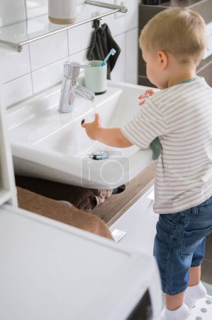 Téléchargez les photos : Vue arrière du petit garçon en vêtements décontractés debout près de l'évier et de la machine à laver tout en se lavant les mains. concept de famille - en image libre de droit