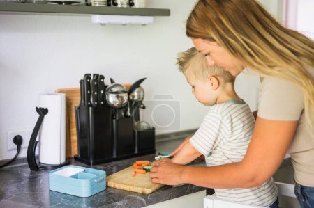 Téléchargez les photos : Vue latérale de la mère gaie en vêtements décontractés debout au comptoir, couper les carottes et les légumes tout en préparant la boîte à lunch avec le petit fils, passer du temps dans la cuisine à la maison. concept de famille - en image libre de droit
