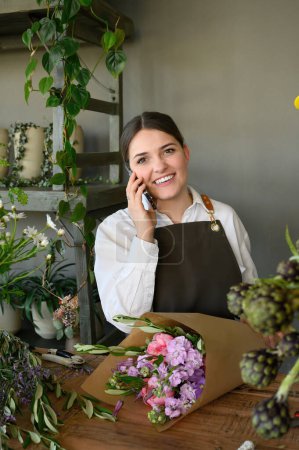 Téléchargez les photos : Fleuriste féminine positive dans un tablier souriant et regardant la caméra tout en parlant sur smartphone et en enveloppant un bouquet de fleurs dans un magasin de fleurs - en image libre de droit