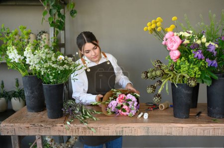 Photo for Focused young female florist in apron standing at table while wrapping bunch of fresh flowers and working in floral shop with potted flowers in daytime - Royalty Free Image