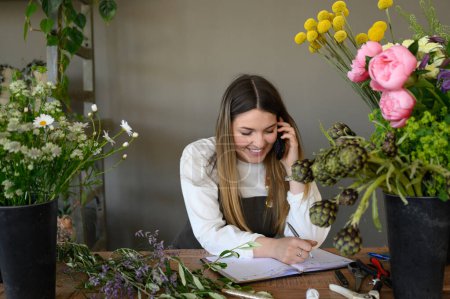 Foto de Joven empresaria sonriente con el pelo rubio escribiendo orden en el cuaderno durante la conversación en el teléfono móvil en la tienda de flores - Imagen libre de derechos