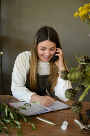 Photo for Positive young female entrepreneur taking orders over cellphone while writing in notebook at table in flower shop - Royalty Free Image