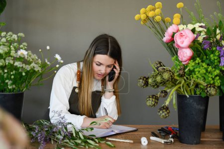 Photo for Young female florist writing client order in notebook during conversation on mobile phone while working at flower shop - Royalty Free Image