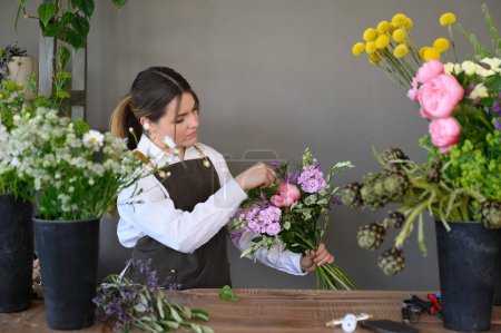 Konzentrierte Dame in Schürze justiert Blumen, während sie im Blumenladen arbeitet und kreative Bouquets herstellt