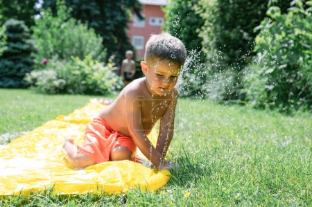 Photo for Side view of adorable little boy sitting on colorful water slide and playing, having fun on green lawn in garden on a summer day. family content, summer content - Royalty Free Image