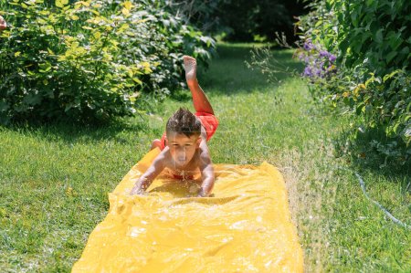 Photo for Little boy jumping on water slide and splashing water in shorts with wet hair on colorful slide in garden and looking at camera, trees and grass in background. family concept - Royalty Free Image