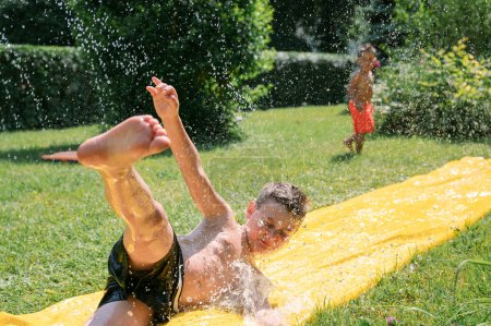 Photo for Kids Having Fun On Water Slide In Garden, sliding on water slide and splashing water, little brother and garden in background.High quality photo. family content - Royalty Free Image