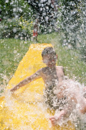 Téléchargez les photos : Vue de face de l'enfant torse nu en short avec les cheveux mouillés avec éclaboussures d'eau dans le jardin par une chaude journée d'été. contenu familial - en image libre de droit