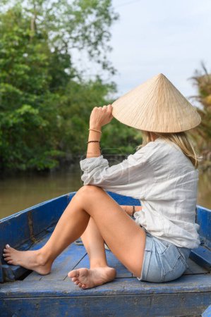 Téléchargez les photos : Vue arrière du caucasien Touriste féminine en vêtements décontractés et chapeau vietnamien assis avec les jambes croisées et regarder la nature sur le bateau pendant le voyage en bateau dans le delta du Mékong, Vietnam - en image libre de droit