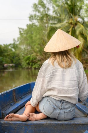 Foto de Vista trasera de caucásica turista femenina en ropa casual, pelo largo y rubio y sombrero vietnamita sentado en barco de madera durante el viaje en barco en el delta del río Mekong, Vietnam - Imagen libre de derechos