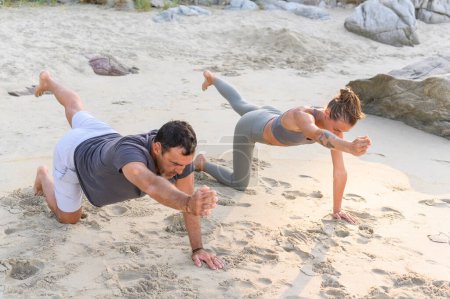 Téléchargez les photos : Vue latérale d'un jeune homme et d'une jeune femme en vêtements de sport faisant des exercices de yoga sur une plage de sable près de la mer au lever du soleil au Mexique - en image libre de droit