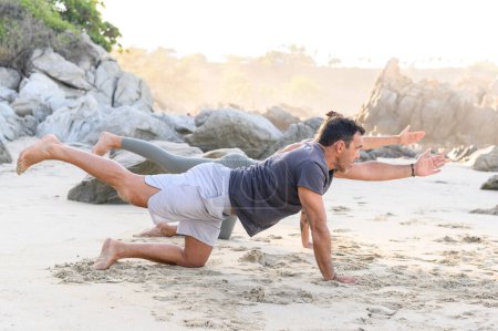 Téléchargez les photos : Vue latérale du jeune homme et de la jeune femme en vêtements de sport faisant du sport sur une plage de sable et étirant les bras tout en pratiquant le yoga par une journée ensoleillée. contenu de style de vie - en image libre de droit