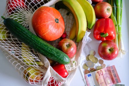 Foto de De composición superior de varias verduras maduras y frutas en bolsa ecológica junto a la factura de supermercado, monedas y dinero sobre fondo blanco - Imagen libre de derechos