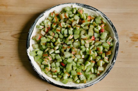 Top view of neatly arranged fresh cut green rhubarb stalk pieces on uncooked pie dough in flat baking pan on wooden table in kitchen in daylight