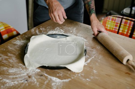 Photo for High angle of anonymous female chef while standing with rolling pin in hand near wooden table with scattered flour and dough arranged neatly on baking pan in daylight - Royalty Free Image