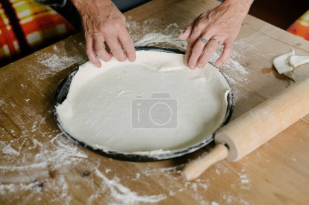 Photo for High angle of anonymous female baker looking down while standing at wooden table with rolling pin scattered flour and cutting extra dough on round baking pan in daylight - Royalty Free Image