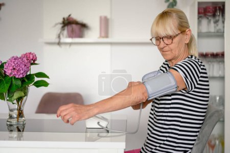 Photo for Side view of focused senior woman in causal clothes and eyeglasses sitting at table and using blood pressure monitor while measuring and checking pulse and blood pressure at home by daylight - Royalty Free Image