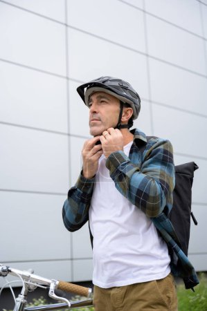 Téléchargez les photos : Homme d'âge moyen en vêtements décontractés avec sac à dos se préparer à monter à vélo mettant casque avant de monter à la maison du bureau. contenu de style de vie - en image libre de droit