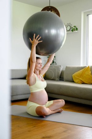 Junge schwangere Frau in Fitnessklamotten sitzt auf Matte und macht zu Hause Sport mit Ball.