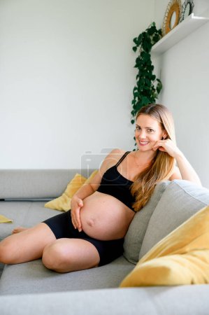 Photo for Front view of cheerful young pregnant female in sporty outfit sitting on sofa, looking at camera, touching belly while resting at home on daytime. - Royalty Free Image