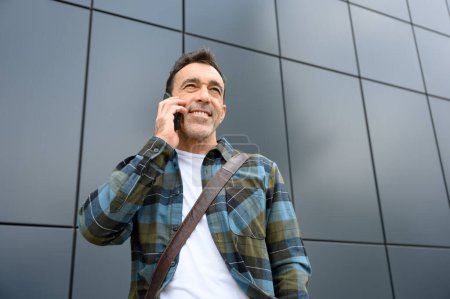 Téléchargez les photos : Homme barbu positif en chemise à carreaux et en jeans parlant sur un téléphone portable et regardant loin tout en se tenant contre un mur gris - en image libre de droit