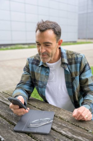 Photo for Young man in casual clothes sitting at wooden table and browsing smartphone - Royalty Free Image