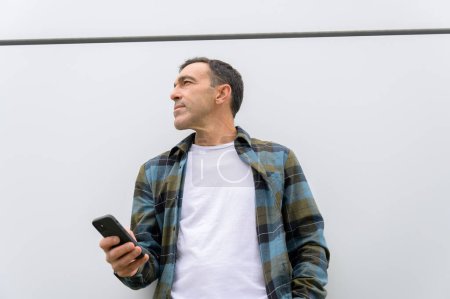 Foto de Bajo ángulo de hombre de mediana edad en camisa a cuadros de pie cerca de la pared gris y el teléfono móvil de navegación mientras mira hacia otro lado. contenido empresarial - Imagen libre de derechos