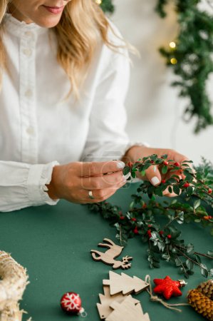Téléchargez les photos : De dessous de la récolte anonyme femelle décorer et tenant couronne de Noël avec des branches vertes avec des baies rouges, fond vert à côté de couronnes différents ornements, trucs d'artisanat. Concept de Noël - en image libre de droit