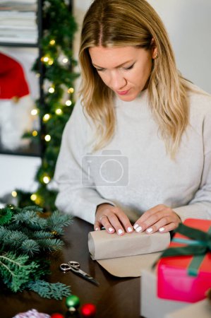 Téléchargez les photos : Jeune femme en pull enveloppant cadeaux de Noël en utilisant du papier, des ciseaux et des rubans colorés à la maison. Préparatifs festifs de vacances. concept de Noël - en image libre de droit