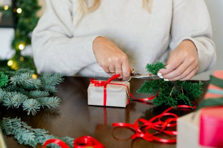 Téléchargez les photos : Récolte de femme méconnaissable en pull enveloppant cadeaux de Noël en papier, en arrière-plan lumières de Noël à côté de son sapin vert et papier d'emballage sur la table - en image libre de droit
