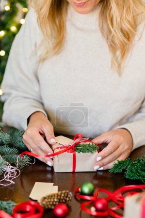 Foto de Cosecha de irreconocible Mujer en jersey con regalo de Navidad decorado con cinta roja y abeto en la mano en la mesa, en el fondo luces de Navidad siguiente. Navidad y Año Nuevo Concepto - Imagen libre de derechos