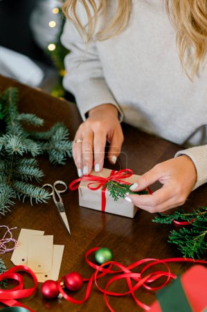 Photo for Crop of woman in sweater wrapping Christmas presents using paper, scissors, colorful ribbons and putting fir on present box at home. In Background lights. Christmas concept - Royalty Free Image