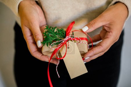 Photo for Crop of unrecognizable Woman in sweater holding christmas present decorated with red ribbon and fir in hand, in background christmas lights next to her green fir and wrapping paper on table - Royalty Free Image