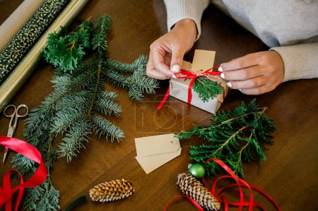 Photo for Crop of womans hand in sweater wrapping Christmas presents using paper, scissors, colorful ribbons and putting fir on present box at home. In Background lights. Christmas concept - Royalty Free Image