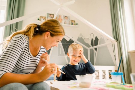 Photo for Front view of blond cute toddler in casual look with mother sitting at table at home and drawing with paints on paper during daytime - Royalty Free Image
