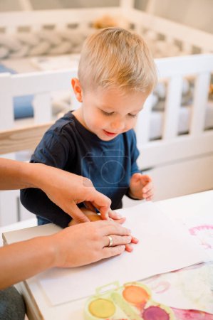 Photo for From above of adorable little boy in casual clothes sitting at table and drawing with colorful paint while spending time together with his mother in cozy room - Royalty Free Image