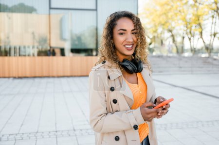 Foto de Mujer joven positiva con auriculares y smartphone sonriendo y mirando a la cámara mientras está de pie en la calle de la ciudad y escuchando música - Imagen libre de derechos
