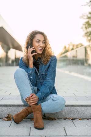 Photo for Full body of cheerful female in denim jacket and jeans speaking on mobile phone while sitting on stairs in city - Royalty Free Image
