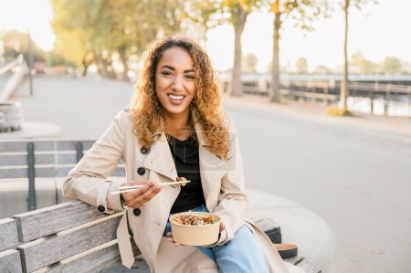 Photo for Happy cheerful young woman sitting, looking at camera and eating asian food bowl with noodles from takeaway paper box using chopsticks city street. Fast food eatery and asian cuisine - Royalty Free Image