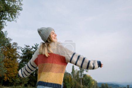 Téléchargez les photos : Du dessous de la jeune femelle en pull chaud et chapeau tricoté debout contre les arbres verts et les nuages tout en profitant d'une journée ensoleillée à la campagne en hiver - en image libre de droit