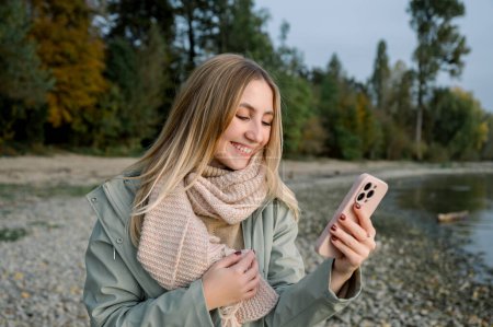 Téléchargez les photos : Positif jeune femme cheveux blonds longs dans des vêtements chauds souriant et naviguant téléphone mobile tout en se tenant sur la rive du lac dans le parc d'automne - en image libre de droit