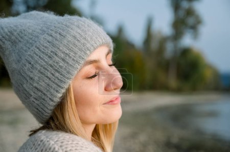 Photo for Side view of young woman in warm cozy sweater and hat, breathes clean air, looks at the sky with a smile and closed eyes, walks with pleasure in the autumn forest. people health content - Royalty Free Image