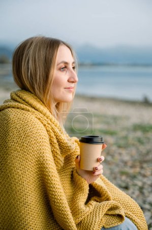 Photo for Outdoor shot of smiling young happy woman freelancer sitting on the ground on blanket and drinking coffee or tea, while having rest in nature near lake and mountains. healthy lifestyle concept - Royalty Free Image
