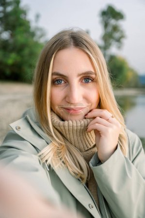 Foto de Mujer joven positiva en abrigo y bufanda mirando a la cámara mientras está de pie sobre el fondo borroso del parque - Imagen libre de derechos