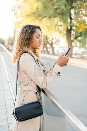 Téléchargez les photos : Jeune femme afro-américaine positive en vêtements décontractés, sac à main noir, debout sur des arbres de fond de rue et des textos ensoleillés sur téléphone portable. concept mode de vie - en image libre de droit