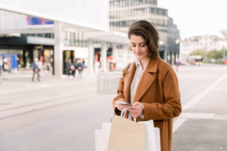 Téléchargez les photos : Jeune femme positive aux cheveux courts en manteau élégant avec sac à main et sacs à provisions regardant un smartphone debout dans la rue de la ville attendant le bus - en image libre de droit