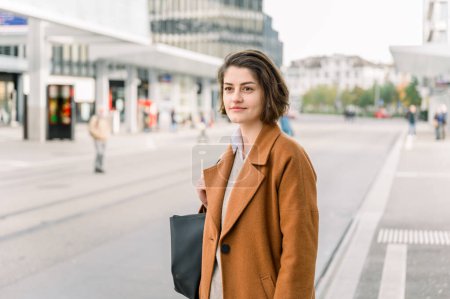 Téléchargez les photos : Vue latérale de la jeune femme aux cheveux courts en manteau élégant avec sac à main debout à la gare routière, en regardant au loin sur le chemin du retour - en image libre de droit